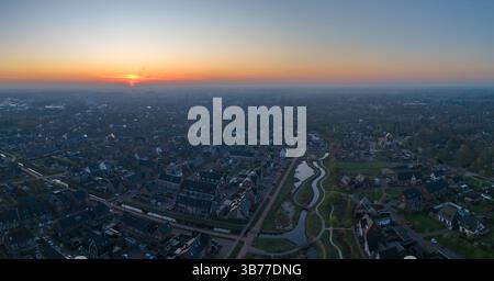 Ein Panoramablick auf den Sonnenaufgang über Nijkerk mit einer ruhigen Wohngegend mit gewundenen Straßen und einem glitzernden Fluss, der das Morgenlicht reflektiert. Stockfoto