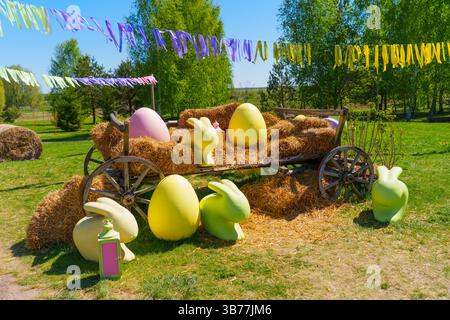 Lebhafte Darstellung großer bunter Ostereier und Hasenfiguren auf einem traditionellen Wagen auf einer sonnigen Wiese, die durch festliche Dekorationen im Hinterteil unterstrichen wird Stockfoto