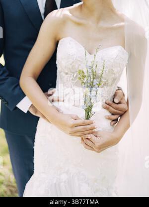 Bräutigam in einem blauen Anzug umarmt von hinten Braut in einem weißen schulterfreien Kleid. Braut hält Wildblumen in den Händen. Stockfoto