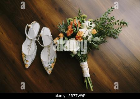 Brautstrauß aus weißen und orangen Rosen, Eringyum, ammi, Buchsbaumzweigen, Alstroemeria mit weißen Bändern und Schuhen der Braut auf dem Boden. Stockfoto
