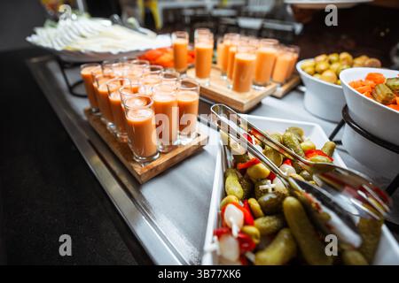Farbenfroher Buffettisch mit eingelegtem Gemüse, frischen Karotten- und Gurkenscheiben und Reihen kleiner Gläser gefüllt mit cremigem Gazpacho oder Gemüse Stockfoto
