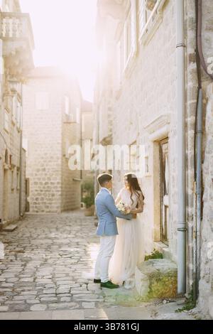 Die Braut und der Bräutigam umarmten sich in einer wunderschönen alten Straße von Perast in der Nähe einer weißen Steinmauer. Stockfoto