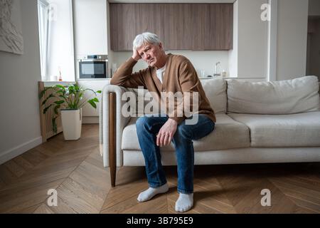 Depressiver, apathischer alter Mann, der Kopf in der Hand ruht und allein auf der Couch sitzt, mit freiem Blick zu Hause Stockfoto