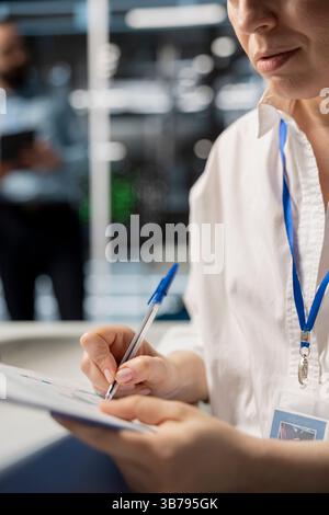 Frau im Rechenzentrum, die sich die Fehlerbehebungen notiert, die nach der Ausführung von Diagnoseskripten auf dem Computer vorgenommen werden müssen. Schreiben der Serverraum-Administratoren Reparaturen, die von Supercomputern nach Auswertung der Leistungskennzahlen benötigt werden Stockfoto