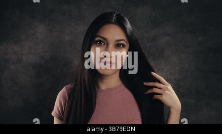 Junge Lateinfrau, die mit ihrem langen, glatten Haar auf dunklem Hintergrund spielt Stockfoto