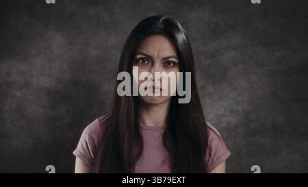 Ernste junge Frau mit langen schwarzen Haaren, die Missbilligung vor dunklem Hintergrund zeigt Stockfoto