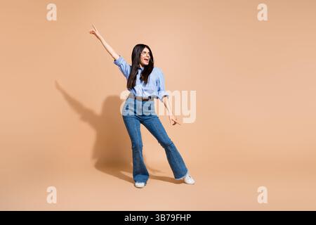 Die junge Frau, die lässig in Jeans und Hemd gekleidet ist, posiert mit ausdrucksstarken Gesten auf beigefarbenem Hintergrund und schafft so eine lebendige Lifestyle-Szene. Stockfoto