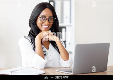 Porträt der schönen schwarzen Geschäftsfrau sitzt an ihrem Schreibtisch mit Laptop, lächelnd und Blick auf die Kamera im Büro Stockfoto