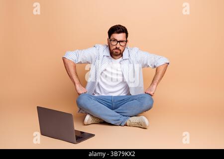 Junger Mann im trendigen Stil, der im Kreuz vor einem Laptop sitzt, auf beigefarbenem Hintergrund und mit entschlossenem Ausdruck gestickt Stockfoto