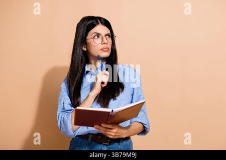 Junge Frau in lässiger Kleidung, die ein Notizbuch hält, während sie tief nachdenkt vor beigefarbenem Hintergrund, die Besinnung und Stil zum Ausdruck bringt. Stockfoto