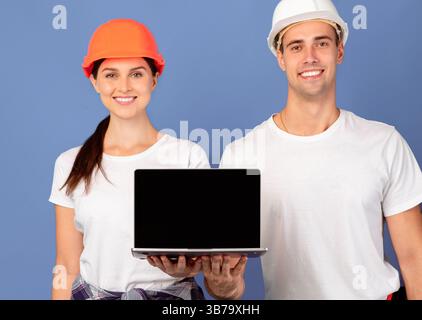 Happy Young Mann Und Frau In Hardhats Holding Laptop Mit Schwarzer Bildschirm Stockfoto