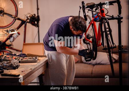 Ein fokussierter Mann in Handschuhen inspiziert ein Fahrradpedal und ist bereit, es an einem roten Fahrrad in einem gemütlichen Innenarbeitsplatz zu befestigen. Stockfoto