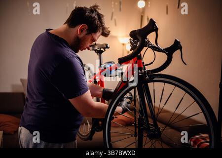 Fokussierter Mann repariert zu Hause ein rotes Rennrad mit Handschuhen und lässiger Kleidung in einem gemütlichen, warm beleuchteten Raum. Stockfoto