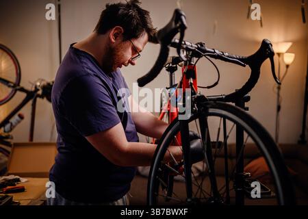 Fokussierter Mann repariert zu Hause ein rotes Rennrad mit Handschuhen und lässiger Kleidung in einem gemütlichen, warm beleuchteten Raum. Stockfoto
