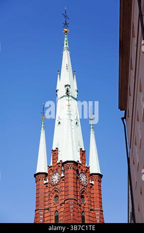 Hohe weiße Kirchtürme mit goldenen Ornamenten erheben sich über einem roten Backsteinturm mit Uhrenscheiben, die vor einem leuchtend blauen Himmel in Stockholm gefangen sind. Stockfoto