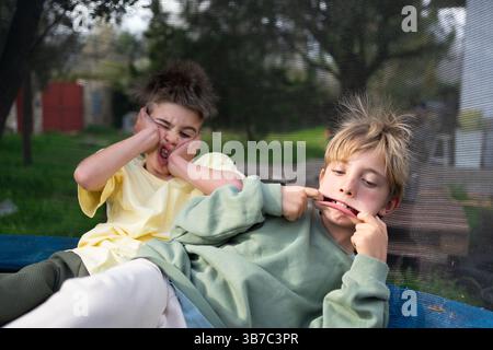 Zwei Brüder ziehen lustige Gesichter, während sie sich entspannen und auf einem Trampolin in ihrem Garten spielen Stockfoto
