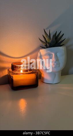 Warmes Ambiente mit Kerzenlicht und gesichtsförmigem Pflanzgefäß, gemütlicher Szene mit beleuchteter Kerze im Glasgefäß und saftiger Pflanze im künstlerischen Topf. Stockfoto
