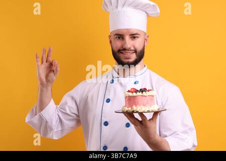 Glücklicher Konditor in Uniform, der Kuchen mit Beeren hält und eine gute Geste auf orangem Hintergrund zeigt Stockfoto