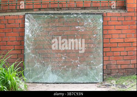 Große Glasscheibe mit Spinnennetzen, die an einer roten Ziegelwand angelehnt sind und bei Tageslicht im Freien mit etwas Grün fotografiert wurden. Stockfoto