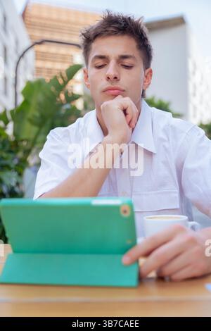 Junger Mann denkt, während er ein Tablet in einem Café im Freien in vertikaler urbaner Umgebung benutzt Stockfoto