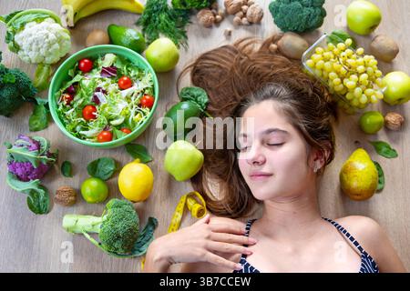 Gesundes Lifestyle-Konzept mit Frau und frischen Produkten. Junge Frau, die sich entspannt, umgeben von frischem Obst, Gemüse und einem grünen Salat, symbolisiert Stockfoto