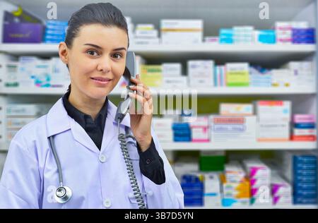 Lächelnde Apothekerin, die mit jemandem am Telefon spricht, während sie einige Bestände im Computer auf Apothekenhintergrund überprüft. Stockfoto