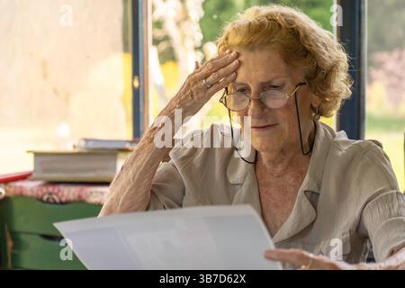 Seniorin trägt Brille, liest einen Brief zu Hause und fühlt eine Welle der Sorge, berührt ihre Stirn mit einer Hand, zeigt eine Mischung von Sorge A Stockfoto