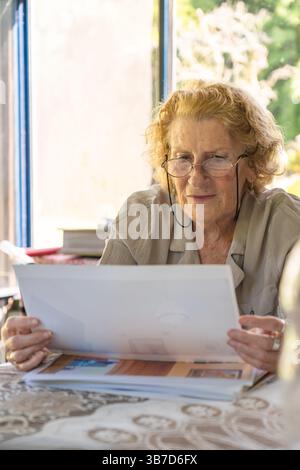 Seniorin mit Brille, überprüft Papiere und Rechnungen, während sie in einem komfortablen häuslichen Umfeld die persönlichen Finanzen verwaltet, wobei Fokus und Stockfoto