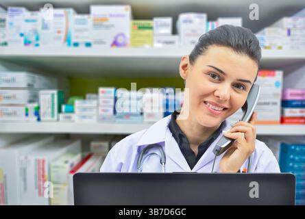 Lächelnde Apothekerin, die mit jemandem am Telefon spricht, während sie einige Bestände im Computer auf Apothekenhintergrund überprüft. Stockfoto