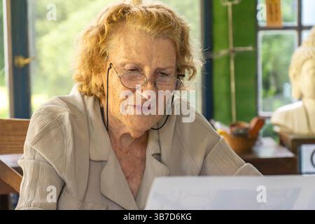Eine ältere Frau mit Brille sitzt an einem Tisch, liest sorgfältig Dokumente mit einem fokussierten Gesichtsausdruck und genießt möglicherweise eine friedliche M Stockfoto