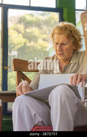 Ältere Frau, die auf einem Holzstuhl in einer Veranda sitzt und Dokumente hält und liest, mit einem ernsten Gesichtsausdruck und natürlichem Licht Stockfoto
