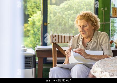 Seniorin sitzt auf einem Stuhl in einer Veranda, hält und liest Dokumente, mit einem Tisch, Stuhl, tragbarer Klimaanlage und einem Fenster mit Gartenblick Stockfoto
