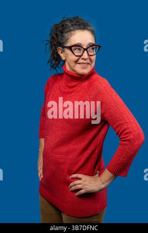 Selbstbewusste reife Frau mit Brille und rotem Rollkragenpullover, die auf einem leuchtend blauen Hintergrund posiert Stockfoto