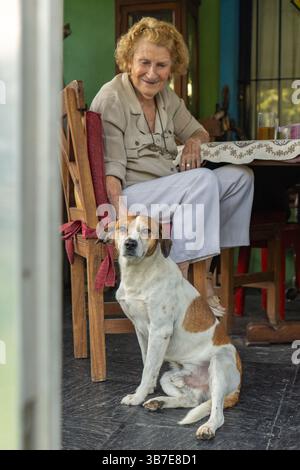 Eine ältere Frau sitzt an einem Tisch in ihrem bunten Haus, streichelt sanft ihren gemischten Hund und teilt einen ruhigen Moment voller Liebe und Gefährten Stockfoto