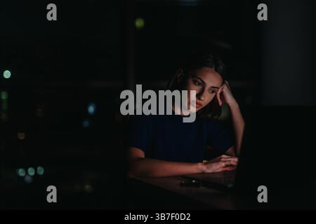 Die junge professionelle Frau konzentriert sich auf ihren Laptop, während sie nach Stunden in einem ruhigen, schwach beleuchteten Büro arbeitet. Stockfoto