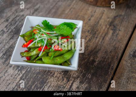 Köstliche Edamame Bohnen mit Chili auf weißer Mini-Schüssel umgeben von hölzernem Hintergrund Stockfoto