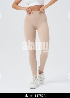 Athletische Frau in beiger Leggings und einem weißen Sport-BH, wobei ihre Hände den Bund anpassen. Sie trägt auch weiße Turnschuhe, stehend Stockfoto