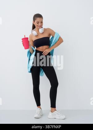 Eine junge Frau mit langen Haaren in sportlicher Kleidung, eine rote Tasse und Kopfhörer, die um ihren Hals liegen, auf einem Fuß stehend, mit schwarzen Leggings Stockfoto