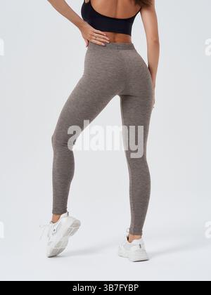 Frau mit grauen Leggings und weißen Turnschuhen, posiert mit Hand auf Hüfte, in einer selbstbewussten Haltung. Schwarzes ärmelloses Crop-Top für sportliche und passgenaue Passform Stockfoto