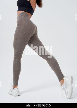 Sportliche Frau in dunklem Sportoberteil und grauer Leggings posiert mit einem ausgestreckten Bein und trägt weiße Sneakers. Das Foto zeigt einen Teil ihres Rückens Stockfoto
