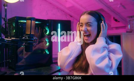 Begeisterte Frau Gamer Mädchen weibliche Streamer in Kopfhörern feiert den Sieg bei Computer Online-Spiel mit fröhlicher Reaktion lächelnd in Neonlicht Stockfoto