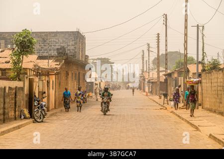 Straßenszene in Ouidah, Benin. Einheimische spazieren und arbeiten inmitten von Gebäuden und tropischen Pflanzen und fangen das authentische tägliche Leben in Westafrika ein. Stockfoto