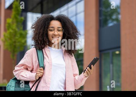 Eine lächelnde junge Frau steht draußen und hält ein Telefon und einen Rucksack. Sie trägt eine Brille und ein pinkfarbenes Hemd. Stockfoto