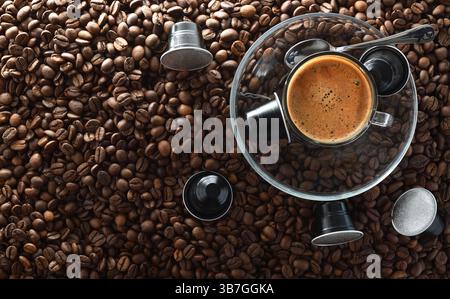 Tasse aufschäumenden Kaffee auf einer Glasplatte in der Mitte auf einem Haufen gerösteter Kaffeebohnen und schwarzer Aluminiumkaffeekapseln. Draufsicht. Stockfoto