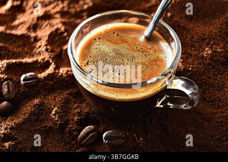 Detail einer Glaskaffeetasse mit frisch gebrühtem Schaumkaffee und einem Metalllöffel auf einem Haufen gemahlenen Kaffees und Bohnen um sie herum. Erhöhte Aussicht. Stockfoto