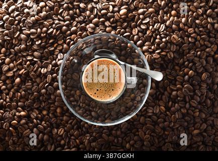 Tasse aufschäumenden Kaffee auf einer Untertasse aus Glas mit einem Metalllöffel in der Mitte auf einem Stapel gerösteter Kaffeebohnen. Draufsicht. Stockfoto