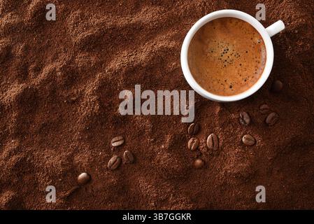 Weiße Kaffeetasse aus Keramik, gefüllt mit schaumigem Kaffee auf einer Seite auf einem Hintergrund aus gemahlenem Kaffee und Bohnen. Draufsicht. Stockfoto
