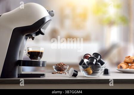 Frisch gebrühter Kaffee in einer Espressomaschine auf einer Holzbank mit Kapseln, Bohnen und Gebäck, mit Wohnzimmer im Hintergrund. Seitenansicht. Stockfoto