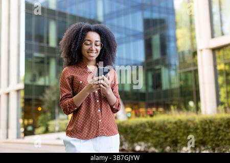 Eine lächelnde afroamerikanerin mit Brille überprüft ihr Smartphone in einer städtischen Umgebung in der Nähe eines Geschäftsgebäudes. Stockfoto