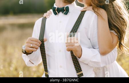 Halbporträt der Braut, die den Bräutigam von hinten umarmt. Braut in einem wunderschönen weißen Kleid. Bräutigam in weißem Hemd, grüner Schleife und farbenfrohen Hosenträgern. Stockfoto